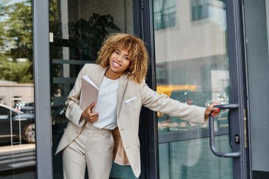 positive african american businesswoman with curly hair holding folder and exiting office building clipart