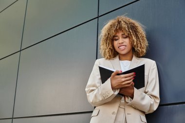Resmi giyimli Afro-Amerikan iş kadını ofis binasının yanındaki defterine bakıyor.