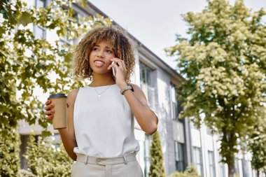 Mutlu Afro-Amerikalı iş kadını telefon görüşmesi yapıyor ve elinde kahve bardağıyla kağıt bardak tutuyor.