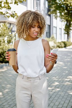 Neşeli Afrikalı Amerikalı iş kadını akıllı telefon kullanıyor ve elinde kahve ile kağıt bardak tutuyor.