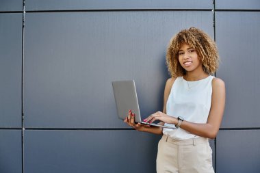 happy curly african american freelancer standing with her laptop near grey wall of office building clipart