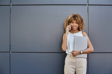 happy young professional, african american woman with laptop making a call against grey backdrop clipart