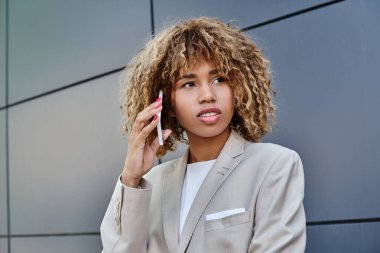 Dalgın ve genç Afrikalı Amerikalı iş kadını kıvırcık saçlı dışarıda akıllı telefondan konuşuyor.