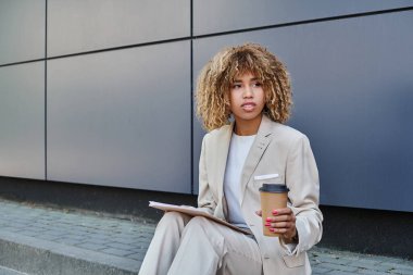 Tarz sahibi, Afro-Amerikan kadın kahve ve klasörle gri ofis duvarının yanında oturuyor.
