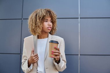 Gri ofis duvarında duran kahve ve klasörlü şık profesyonel Afro-Amerikan kadın.