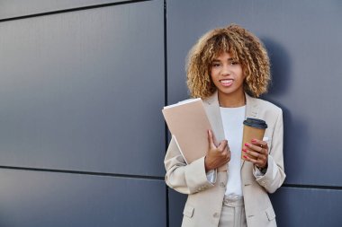 Profesyonel vesikalık fotoğraf, mutlu Afro-Amerikan kadın kahve ve klasörle ofis duvarının yanında duruyor.