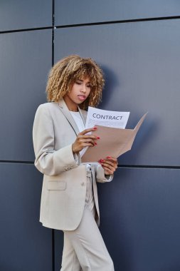 Genç Afro-Amerikan kıvırcık iş kadını Gri ofis duvarının yanındaki belgeyi inceliyor.