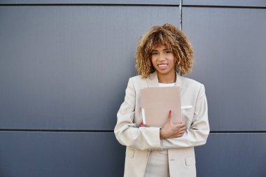 Resmi olarak Afro-Amerikalı genç bir iş kadını gri ofis duvarının yanında klasör tutuyor.