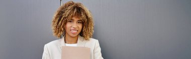Pozitif genç Afrikalı Amerikalı iş kadını resmi olarak ofis duvarının yanında klasör taşıyor.