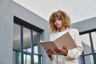 focused african american curly businesswoman reviewing paperwork in folder near office building clipart