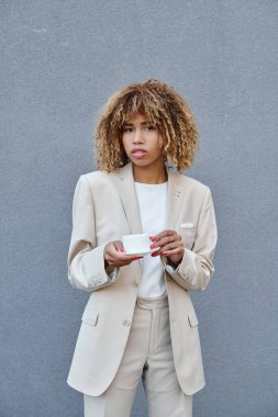 curly african american businesswoman posing with cup of coffee near grey wall of office building clipart