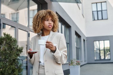 Kıvırcık Afrikalı Amerikalı iş kadını elinde bir fincan kahveyle ofis girişinin yanında duruyor.