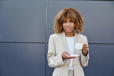 Neşeli Afro-Amerikan iş kadını elinde bir fincan kahveyle ofis binasının girişinde duruyor.