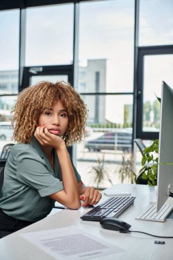 Kıvırcık genç Afrikalı Amerikalı iş kadını ofiste bilgisayar monitörünün önünde oturuyor.