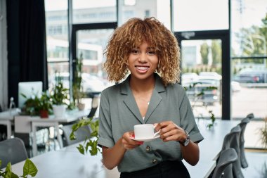 happy african american businesswoman smiling and enjoying coffee break in contemporary office clipart