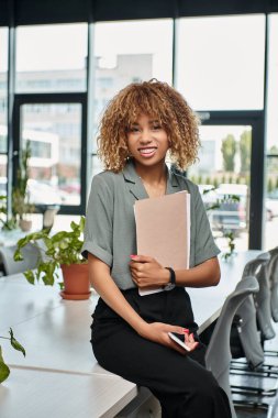 Ofiste kağıt klasörü ve akıllı telefonu tutarken gülümseyen Afro-Amerikan iş kadını.