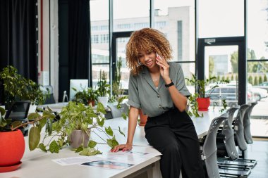 Ofis masasındaki çizelgelere bakarak telefon görüşmesi yapan neşeli Afro-Amerikan iş kadını.