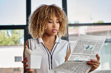 Kıvırcık Afrikalı Amerikalı kadın, karton kutuda Asya yemeği yiyor ve gazete okuyor.