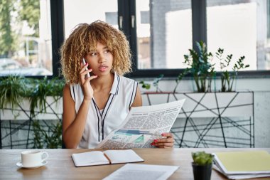 Kıvırcık Afrikalı Amerikalı iş kadını akıllı telefonla konuşuyor ve ofisinde gazetesini tutuyor.