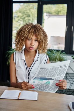 Kıvırcık Afrikalı Amerikalı iş kadını elinde gazete tutuyor ve masa başında oturuyor.