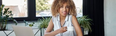 Mutlu Afro-Amerikan iş kadını diş telleriyle elinde bir fincan kahveyle dizüstü bilgisayar ve afiş taşıyor.