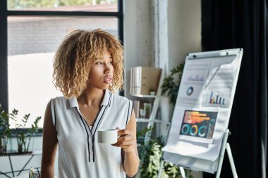 curly african american businesswoman with braces holding cup of coffee beside charts and graphs clipart