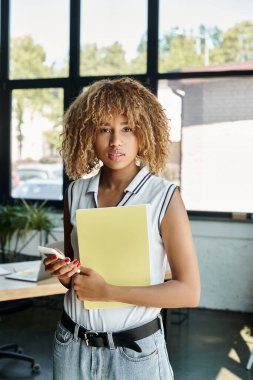 Kıvırcık Afrikalı Amerikalı girişimci diş teli takıyor, elinde belgeler ve akıllı telefon var.