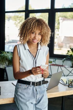 Kıvırcık ve mutlu Afro-Amerikan iş kadını akıllı telefonlu, güneşli ofiste bağlı kalıyor.