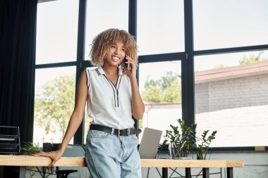 curly and happy african american businesswoman having phone call in sunlit modern office clipart