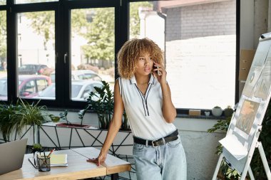 curly and pensive african american businesswoman having phone call in sunlit modern office clipart