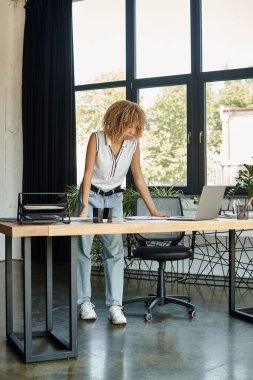 Odaklanmış genç Afrikalı Amerikalı kadın masasında duruyor ve iş günü boyunca dizüstü bilgisayarına bakıyor.