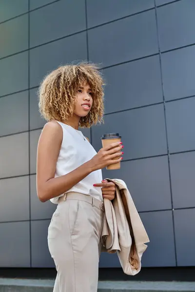 stock image Stylish and curly african american businesswoman with coffee standing against a modern building