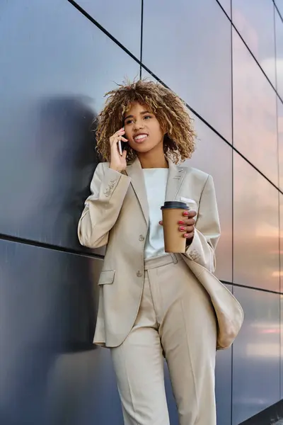 stock image happy african american businesswoman standing with coffee and talking by phone against grey wall