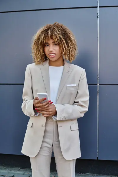 stock image happy african american businesswoman with curly hair standing in suit and using her smartphone