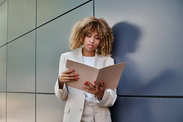 stock image focused african american curly businesswoman reviewing paperwork in folder near grey office wall