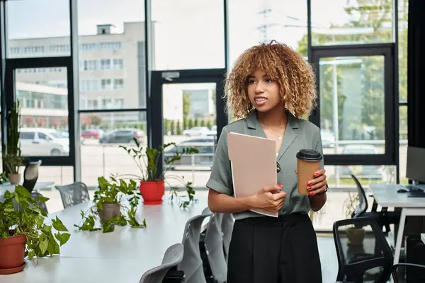 stock image curly african american businesswoman in her 20s, standing with coffee and folder in office