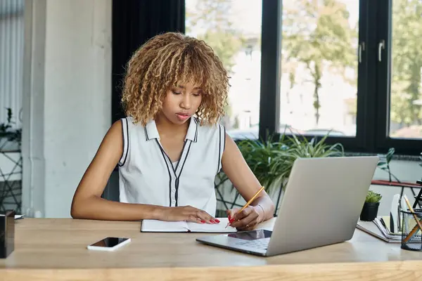 集中精神的非洲裔美国女商人在一个时髦的办公室里 在笔记本电脑旁边做笔记 — 图库照片