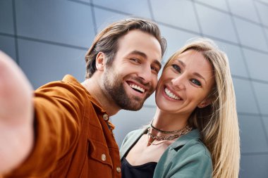 A stylish man and woman posing together, taking a selfie by a modern building. clipart