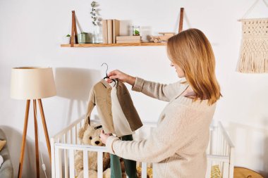 young future mother holding baby clothes near crib with soft toys in nursery room, birth expectation clipart