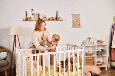 Neşeli genç bir anne, bebek beşiğinin yanında tatlı bir çocuğu kucağında tutuyor, mutlu bir annelik.