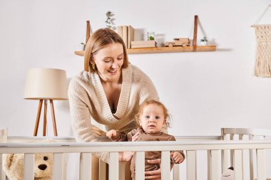 smiling woman embracing adorable son standing in crib in nursery room at home, care and support clipart