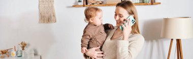 smiling woman with toddler son in hands talking on mobile phone in nursery room, horizontal banner clipart