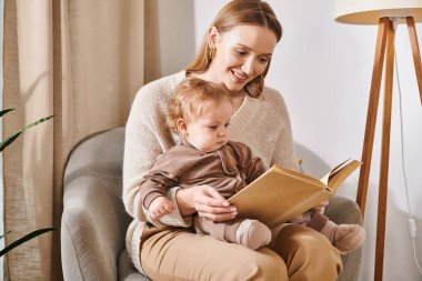 Mutlu anne, sevimli bebeğe kitap okuyor rahat çocuk odasında koltukta oturuyor, modern ebeveynlik yapıyor.