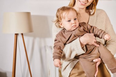 young smiling woman holding and embracing toddler son in cozy nursery room, blissful motherhood clipart