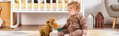 cute little boy playing with toy horse near crib on floor in nursery room, horizontal banner clipart