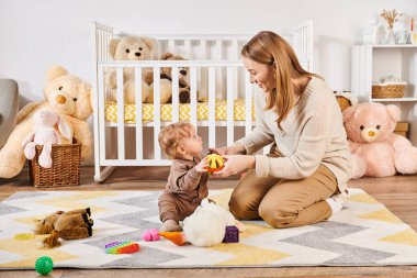 Bebek odasında bebek beşiğinin yanında yumuşak oyuncaklarla oynayan mutlu anne ve çocuk, modern ebeveynlik