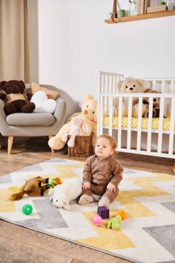 Tatlı küçük çocuk yumuşak oyuncakların yanında yerde oturuyor ve bebek odasında rahat bir beşik, mutlu bir çocukluk.