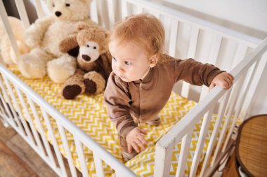 high angle view of toddler boy standing in crib with soft toys in nursery room at home, childhood clipart