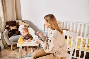 Genç bir kadın, bebeğini bebek koltuğunda kahvaltıyla besliyor, çocuk odası, modern ebeveynlik...