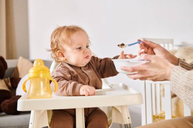 Anne, tapılası oğlunu bebek sandalyesinde kahvaltıyla besliyor. Çocuk odası, modern ebeveynlik.